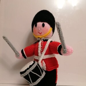 May include: A knitted toy soldier drummer in a red jacket with white trim, black pants, and a black hat. The toy has a pink face, a yellow collar, and holds gray drumsticks. A white and silver drum hangs from a white strap.