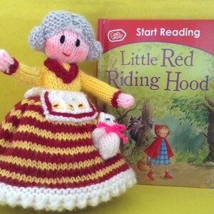 May include: A handmade knitted doll of a grandmother figure, wearing a yellow top, red and white striped skirt, and a white apron. Beside the doll is a book titled "Little Red Riding Hood". The book cover features an illustration of the story.