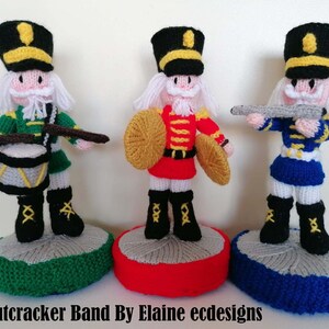 May include: Three handmade knitted nutcracker figures in a band. The figures are dressed in green, red, and blue uniforms, each with a black hat and white beard. One figure holds drumsticks and a drum, another a shield, and the third a rifle. The Nutcracker Band By Elaine ecdesigns.