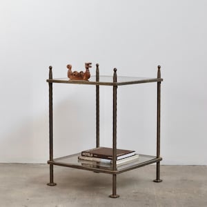 May include: A square two-tier side table with a dark bronze-toned metal frame and glass shelves. A small wooden dragon figurine sits on the top shelf, while two books titled Hotels and Fireplaces are stacked on the bottom shelf.