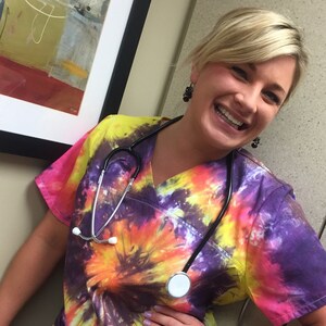May include: A tie-dye scrub top in shades of yellow, orange, pink, and purple. A stethoscope is draped around the neck. The top has a V-neck and pockets. The person is smiling.