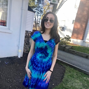May include: A woman wearing a blue and green tie-dye dress with a knotted neckline. She is wearing sunglasses and has long brown hair.