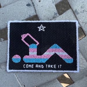 May include: Black cross-stitch art piece with a white border. The design features a stylized figure in pink, blue, and white, with a star and the text "COME AND TAKE IT".