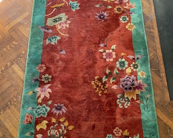 Op de afbeelding: Een vintage Chinees tapijt met een rode achtergrond en een turquoise rand. Het tapijt heeft een bloemenpatroon in verschillende kleuren, waaronder roze, paars, geel en groen.