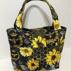 May include: Black tote bag with a sunflower and bee pattern. The bag has two handles and a black lining. The pattern is on a black background with gold accents.