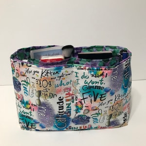 May include: A white fabric tote bag with multiple pockets. The fabric has a colorful, abstract print with words like "Gimme Five", "Are you Kitten", "I do what I want", and "Attitude".