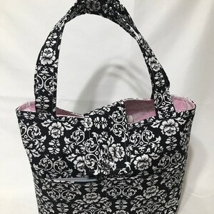 May include: Black and white tote bag with a floral pattern. The bag has a pink lining and a zippered pocket on the front.