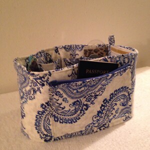 May include: A white and blue paisley patterned fabric organizer with multiple pockets and a zippered pocket. The organizer is filled with items such as a passport, sunglasses, and a small pouch.