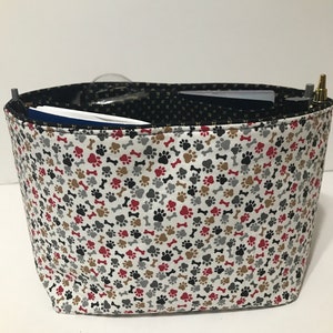 May include: A white fabric tote bag with a black, red, brown, and gray paw print and bone pattern. The bag is lined with black fabric and has a gold zipper.