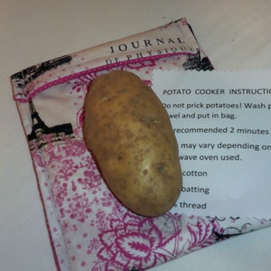 May include: A single potato sits on a pink and white fabric bag with a black Eiffel Tower design. The bag has the text "JOURNAL DE PHYSIQUE" on it. A white piece of paper with the text "POTATO COOKER INSTRUCTION: Do not prick potatoes! Wash potatoes and towel and put in bag. recommended 2 minutes at a time until potatoes are cooked. Time may vary depending on the model microwave oven used. cotton batting 6 thread" is partially visible.