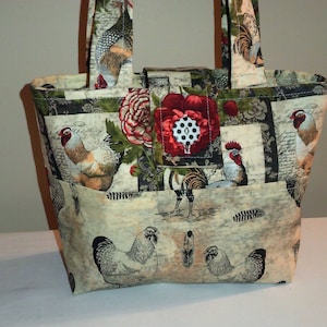 May include: A beige tote bag with a patchwork design featuring roosters, flowers, and feathers. The bag has a red flower with a white polka dot button on the front.