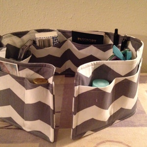 May include: A gray and white chevron patterned fabric organizer with multiple pockets. The organizer is filled with various items, including a passport, pens, and a small bottle.