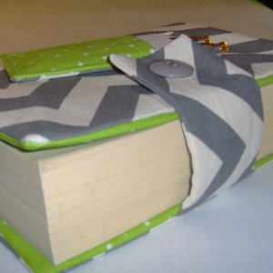 May include: A gray and white chevron book cover with a green polka dot bookmark and a gray fabric strap with a button. The book cover is made of fabric and has a green trim.