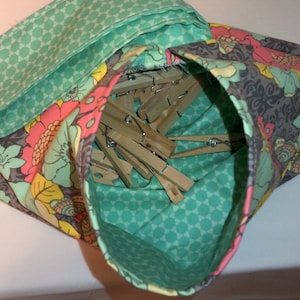 Könnte beinhalten: Eine grüne Wäschetasche aus Stoff mit floralem Muster und Kordelzug. Die Tasche ist mit Holz Wäscheklammern gefüllt.