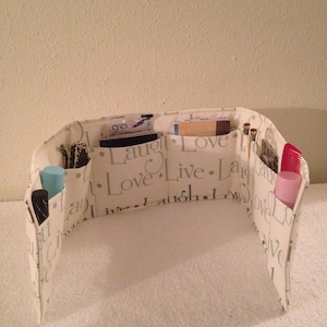 May include: A white fabric organizer with multiple pockets and compartments. The organizer has the words "Laugh. Love. Live. Laugh. Love. Live." printed on it. The organizer is filled with various items, including makeup, pens, and a small notebook.