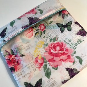 Puede incluir: Una bolsa de tela blanca con estampado floral que presenta rosas rosas, hojas verdes y flores moradas. La bolsa tiene un ribete azul turquesa y un diseño de mariposa negra. La tela tiene un estampado de texto vintage con las palabras "17th, COURT" y "THCT YOU".