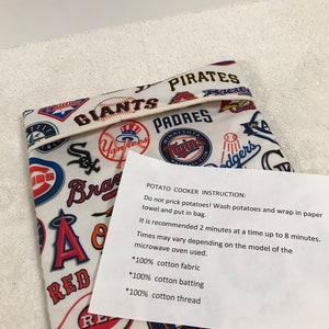 Op de afbeelding: Een witte stoffen tas met verschillende baseballteamlogo's erop gedrukt. De tas is dubbelgevouwen en heeft een wit label met de tekst "POTATO COOKER INSTRUCTION: Do not prick potatoes! Wash potatoes and wrap in paper towel and put in bag. It is recommended 2 minutes at a time up to 8 minutes. Times may vary depending on the model of the microwave oven used. *100% cotton fabric *100% cotton batting *100% cotton thread".