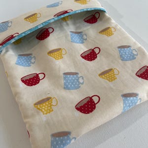 May include: A beige fabric pouch with a blue and white trim. The pouch is decorated with a repeating pattern of red, yellow, and blue teacups.