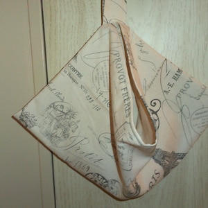 Könnte beinhalten: Eine beige Stofftasche mit einem schwarz-weißen Druck aus französischem Text und Bildern. Die Tasche hat einen langen Riemen und ist in der Mitte gefaltet.