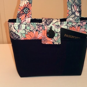 May include: A black tote bag with a floral print and a black button closure. The bag has a pocket on the front that says "PASSPORT".