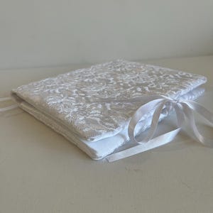 Può includere: Un cuscino portafedi quadrato bianco con un rivestimento in pizzo floreale. Il cuscino è legato con un nastro di raso bianco. Il cuscino viene probabilmente utilizzato per una cerimonia nuziale.