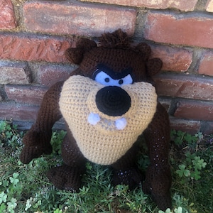 May include: A handmade crochet Taz plush toy. The plush features a brown body, a tan face, and a black nose. The eyes are white with black accents, and the mouth has two white pom-poms. The plush is sitting on green grass in front of a brick wall.