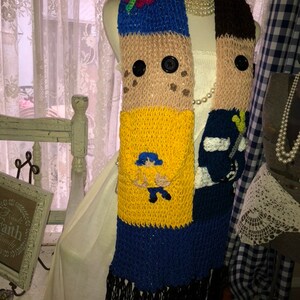 May include: A knitted scarf with a colorful patchwork design featuring a cartoon character, a butterfly, and buttons. The scarf is made of brown, yellow, blue, and black yarn.