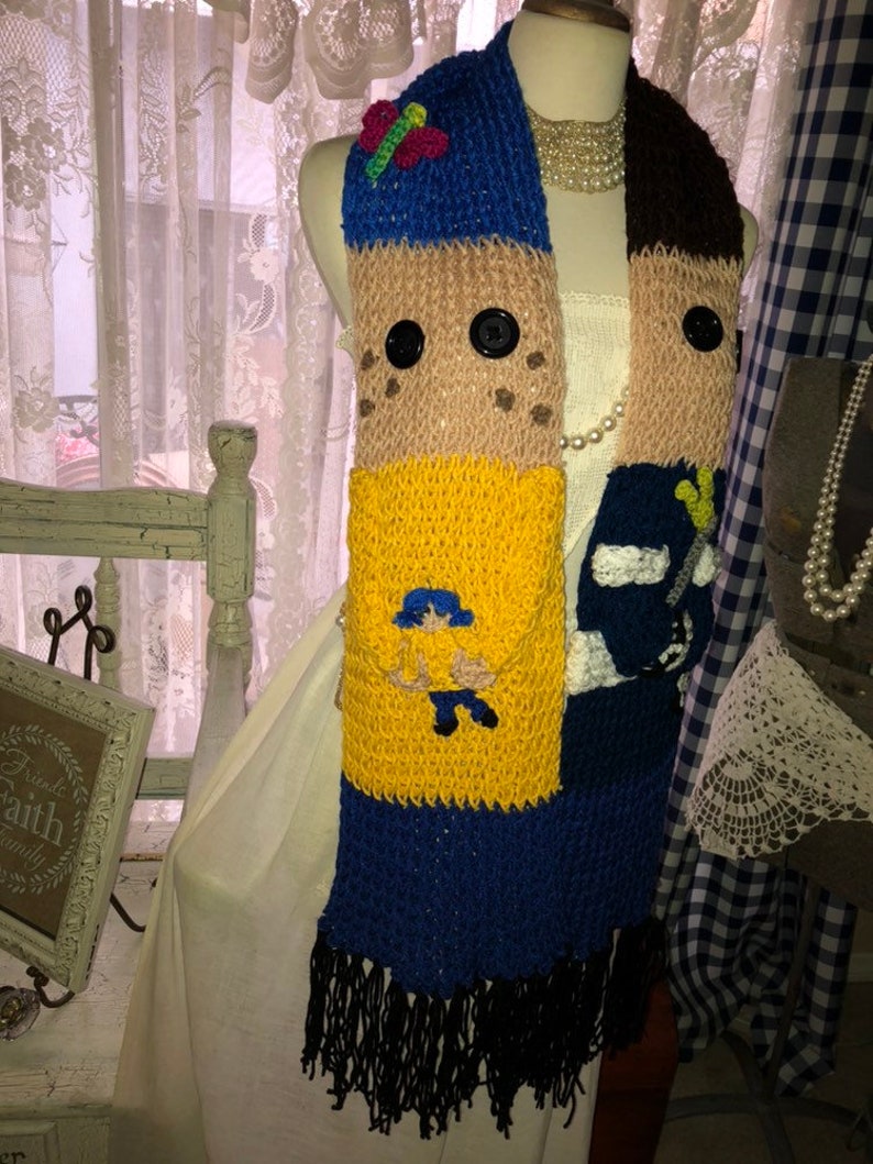 May include: A knitted scarf with a colorful patchwork design featuring a cartoon character, a butterfly, and buttons. The scarf is made of brown, yellow, blue, and black yarn.
