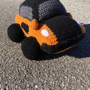 May include: A crocheted toy car in black, gray, and orange. The car has a black roof and a gray windshield. The car is sitting on a gray surface.