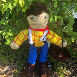 May include: Crocheted toy figure of a cowboy with brown hair, a yellow and red plaid shirt, blue jeans, and brown boots. The toy is holding a brown hat.