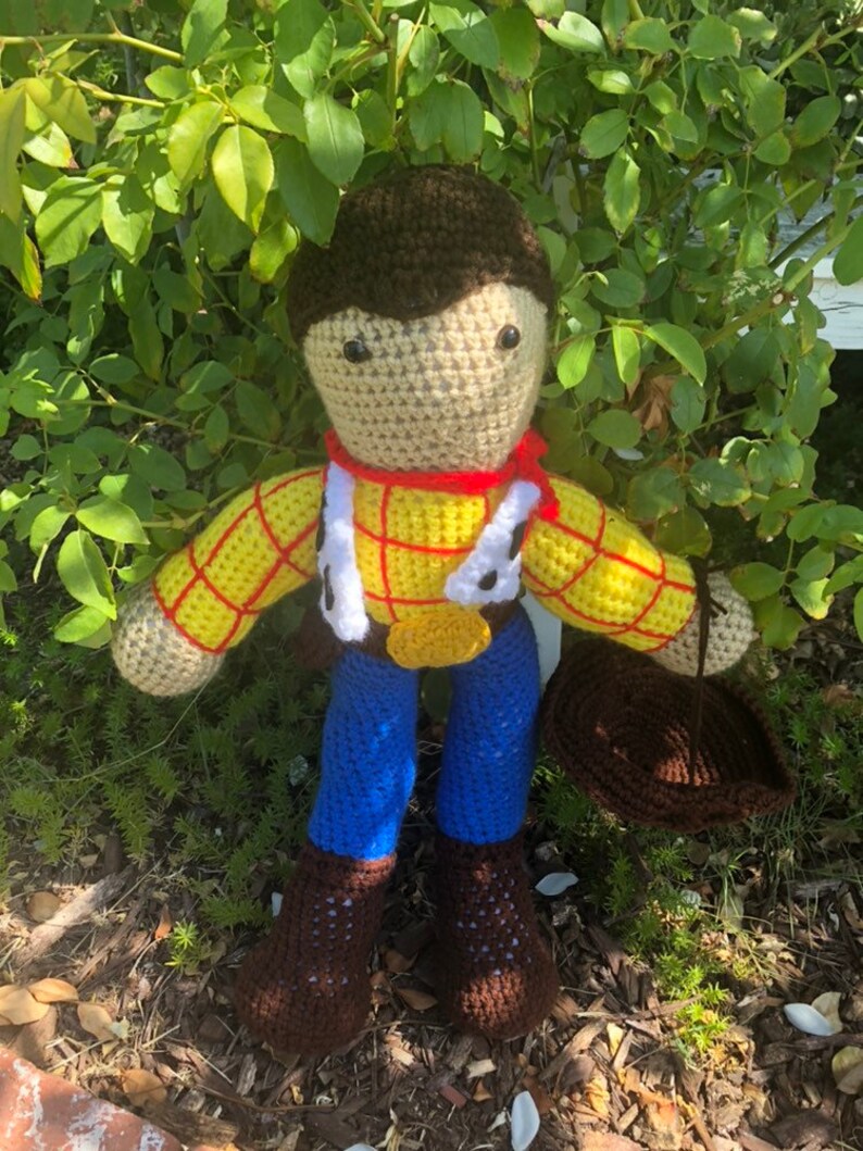 May include: Crocheted toy figure of a cowboy with brown hair, a yellow and red plaid shirt, blue jeans, and brown boots. The toy is holding a brown hat.