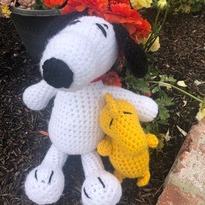 Op de afbeelding: Witte gehaakte Snoopy pluche met zwarte ogen en neus, die een gele gehaakte pluche vasthoudt. De speelgoedjes liggen op een bed van bruine mulch.