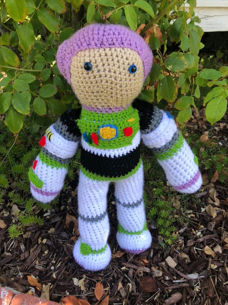 May include: A crocheted Buzz Lightyear toy with a purple helmet, green and white spacesuit, and black and white stripes on the arms and legs. The toy has blue eyes and is standing on a bed of wood chips.