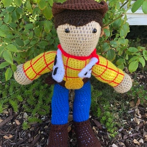 May include: Crocheted plush toy of Woody from Toy Story. The toy is wearing a brown cowboy hat, a yellow and red plaid shirt, a blue vest, blue jeans, and brown boots.