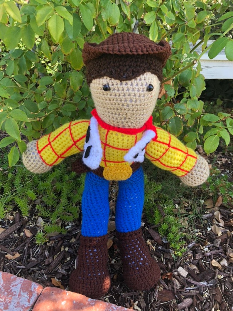 May include: Crocheted plush toy of Woody from Toy Story. The toy is wearing a brown cowboy hat, a yellow and red plaid shirt, a blue vest, blue jeans, and brown boots.