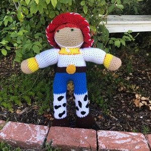 May include: Crocheted plush toy of a cowboy character with a red hat, white shirt, blue jeans, and brown boots. The toy is standing on a brick path.