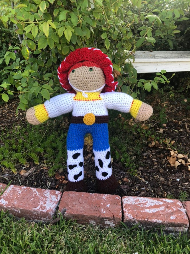 May include: Crocheted plush toy of a cowboy character with a red hat, white shirt, blue jeans, and brown boots. The toy is standing on a brick path.