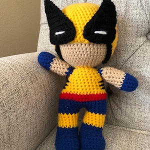 May include: A handmade crochet doll of a superhero character. The doll features a yellow head with black mask and ears, a tan face, and a yellow and blue body with red accents. The arms and legs are blue and tan.