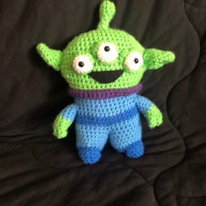 Op de afbeelding: Een groen gehaakt alien speelgoed met drie ogen, een paarse kraag en blauw-groene gestreepte benen.