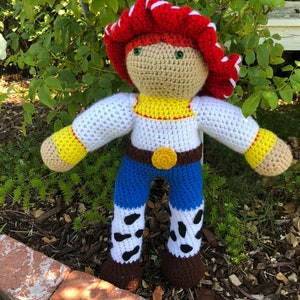 May include: Crocheted plush toy of a character from the Toy Story movie franchise. The toy is wearing a red and white cowboy hat, a white shirt with a yellow vest, blue jeans with white and black spots, and brown boots.