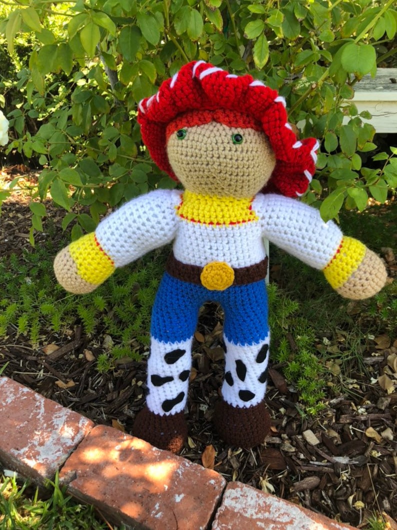 May include: Crocheted plush toy of a character from the Toy Story movie franchise. The toy is wearing a red and white cowboy hat, a white shirt with a yellow vest, blue jeans with white and black spots, and brown boots.