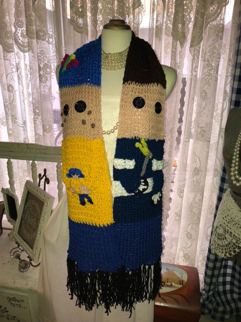 May include: A crocheted scarf with a patchwork design featuring two cartoon faces. The scarf is made of blue, yellow, brown, and white yarn and has black fringe.