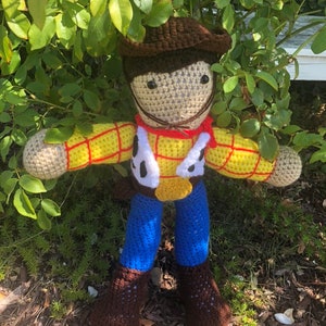 May include: A crocheted toy figure of Woody from the Toy Story franchise. The figure is wearing a brown cowboy hat, a yellow and red plaid shirt, a brown belt, blue jeans, and brown boots. The figure is standing in front of a green bush.