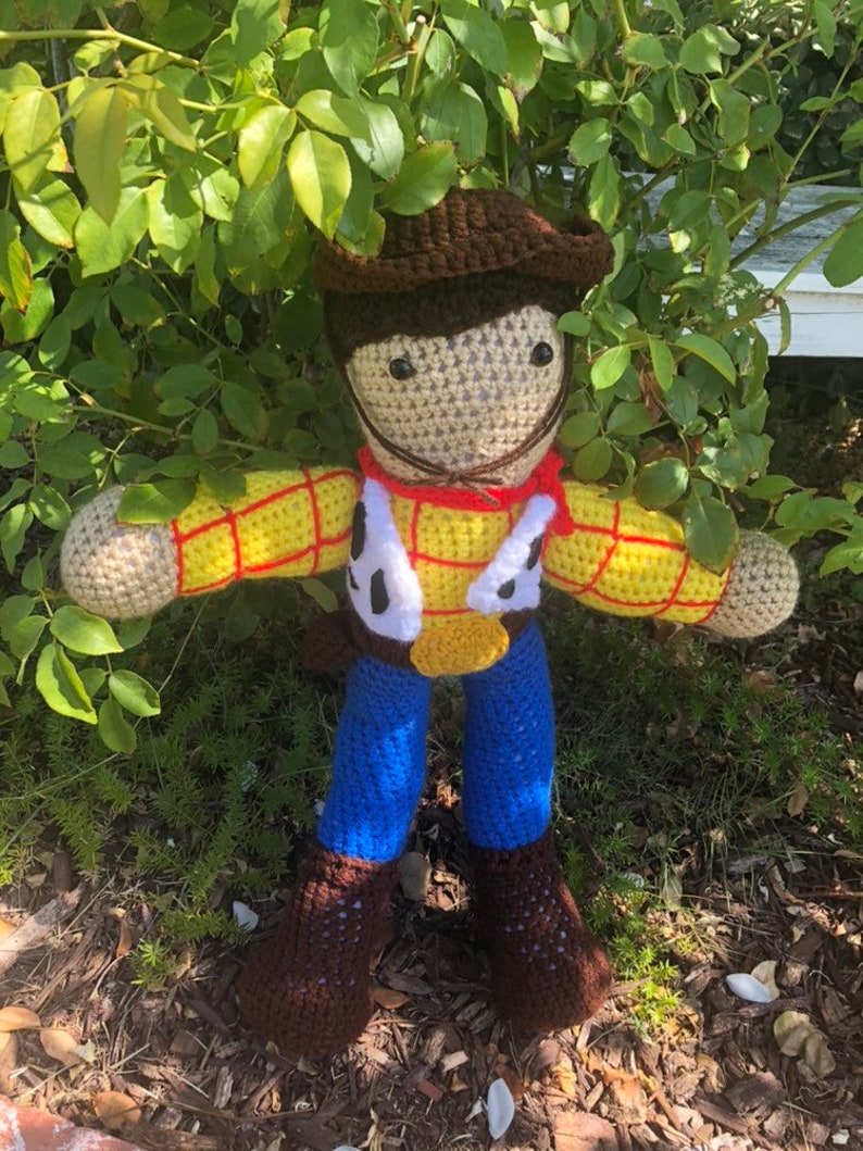 May include: A crocheted toy figure of Woody from the Toy Story franchise. The figure is wearing a brown cowboy hat, a yellow and red plaid shirt, a brown belt, blue jeans, and brown boots. The figure is standing in front of a green bush.