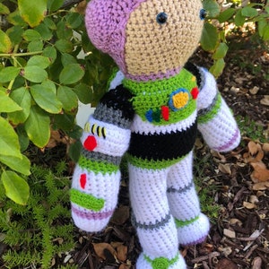 May include: A crocheted Buzz Lightyear toy with purple, white, green, and black details. The toy has a purple helmet and blue eyes.
