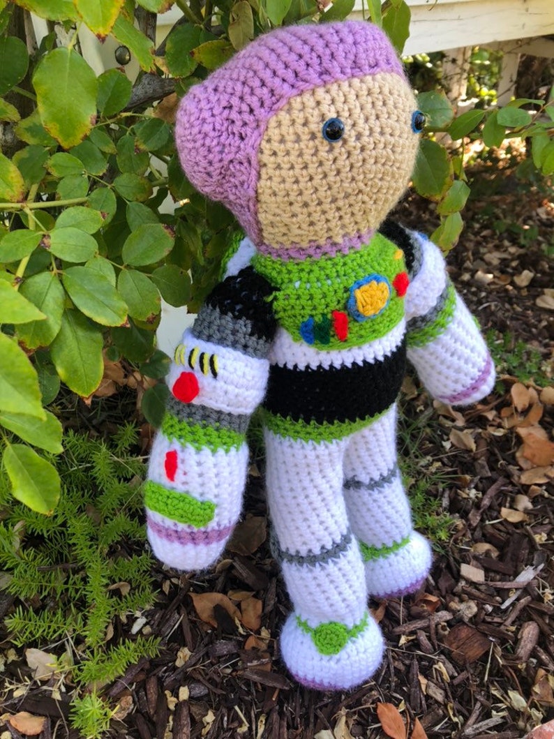 May include: A crocheted Buzz Lightyear toy with purple, white, green, and black details. The toy has a purple helmet and blue eyes.