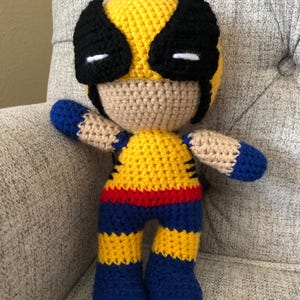 Op de afbeelding: Een handgemaakte gehaakte pop die Wolverine voorstelt. De pop heeft een geel en zwart masker, een geel en blauw kostuum en een bruine huid. De pop is ongeveer 25 cm hoog en is gemaakt van garen. Een uniek, handgemaakt item.
