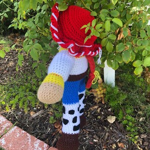 May include: A crocheted toy figure of Woody from the Toy Story franchise. The toy is wearing a red cowboy hat, a white shirt, a blue vest, and brown boots. The toy is standing in a garden setting.