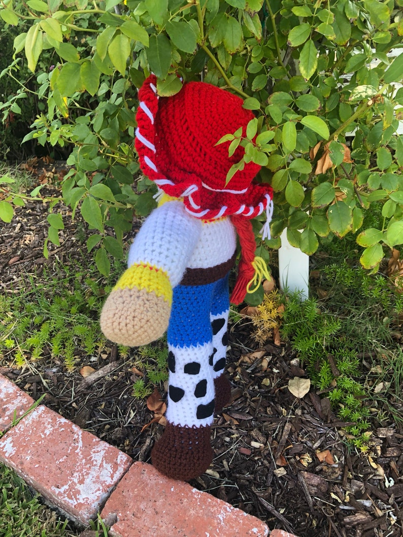May include: A crocheted toy figure of Woody from the Toy Story franchise. The toy is wearing a red cowboy hat, a white shirt, a blue vest, and brown boots. The toy is standing in a garden setting.