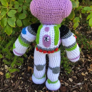 May include: A crocheted plush toy of Buzz Lightyear from the Toy Story franchise. The toy is purple, white, green, and gray. The toy is standing on a bed of mulch.